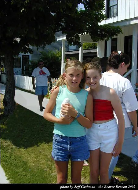 Two girls smile together outdoors beside a house, with other people, trees, and a sidewalk behind them.