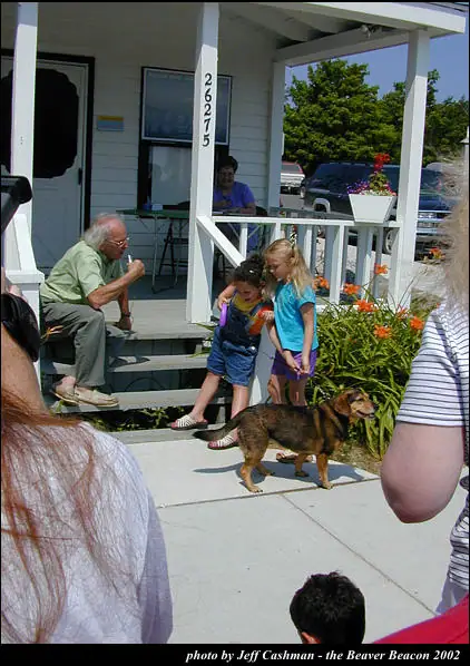 An older man sits on porch steps as two girls stand beside a dog and flowering plants.