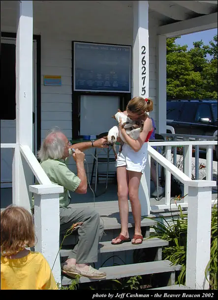 A woman holds a small white dog while an older man with a microphone sits on porch steps.
