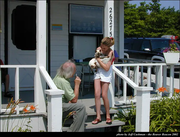 An older man with a microphone speaks to a woman holding a cat on a flower-lined porch.