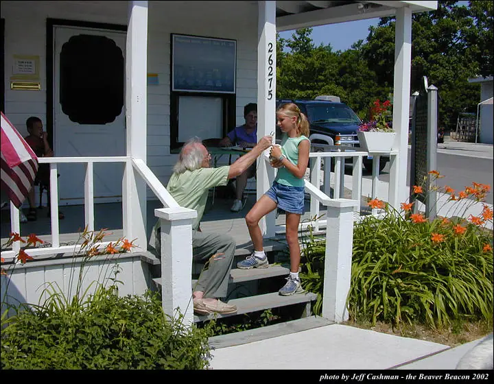 An older man on porch steps hands a small object to a girl beside flower beds.