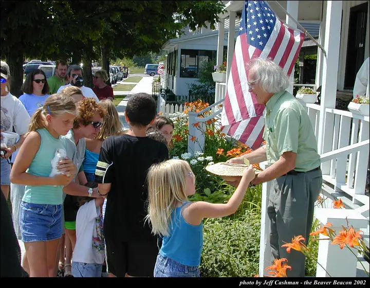 An older man hands a woven basket to a girl as a crowd gathers beside a flower-filled porch.