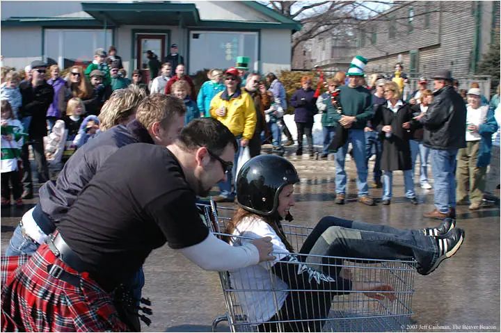A man in a tartan kilt pushes a shopping cart carrying a helmeted woman before a watching crowd.