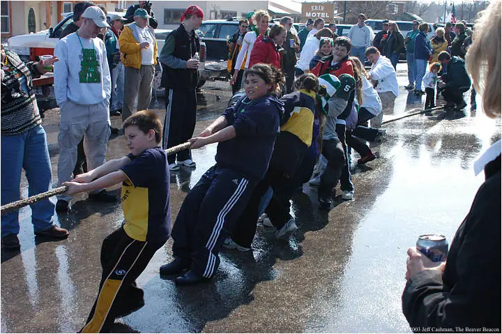 Children pull in a tug-of-war on a wet street while spectators watch.