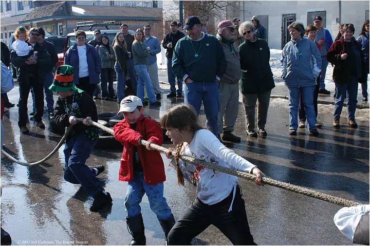 Children pull a thick rope across wet pavement as onlookers gather around.