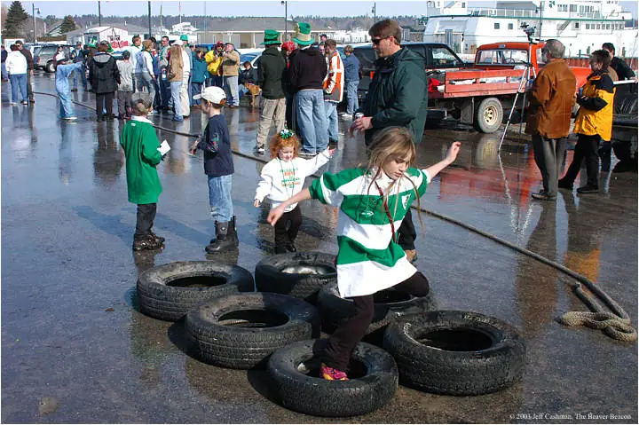 Children navigate a tire obstacle course on wet pavement as spectators gather beside a large white vessel.