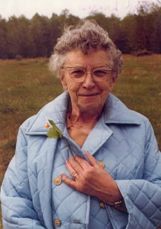 Portrait of an elderly woman wearing glasses and a blue quilted jacket, with an orange flower pinned near her collar.