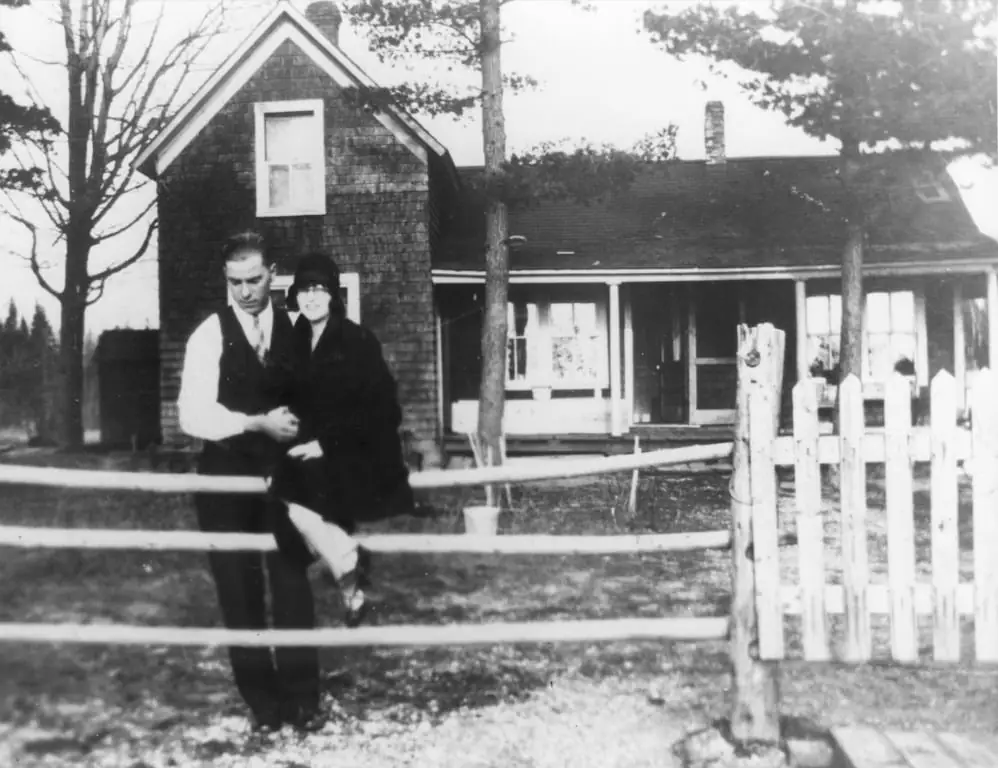 Two people stand behind a wooden fence in front of a shingled house with a porch.