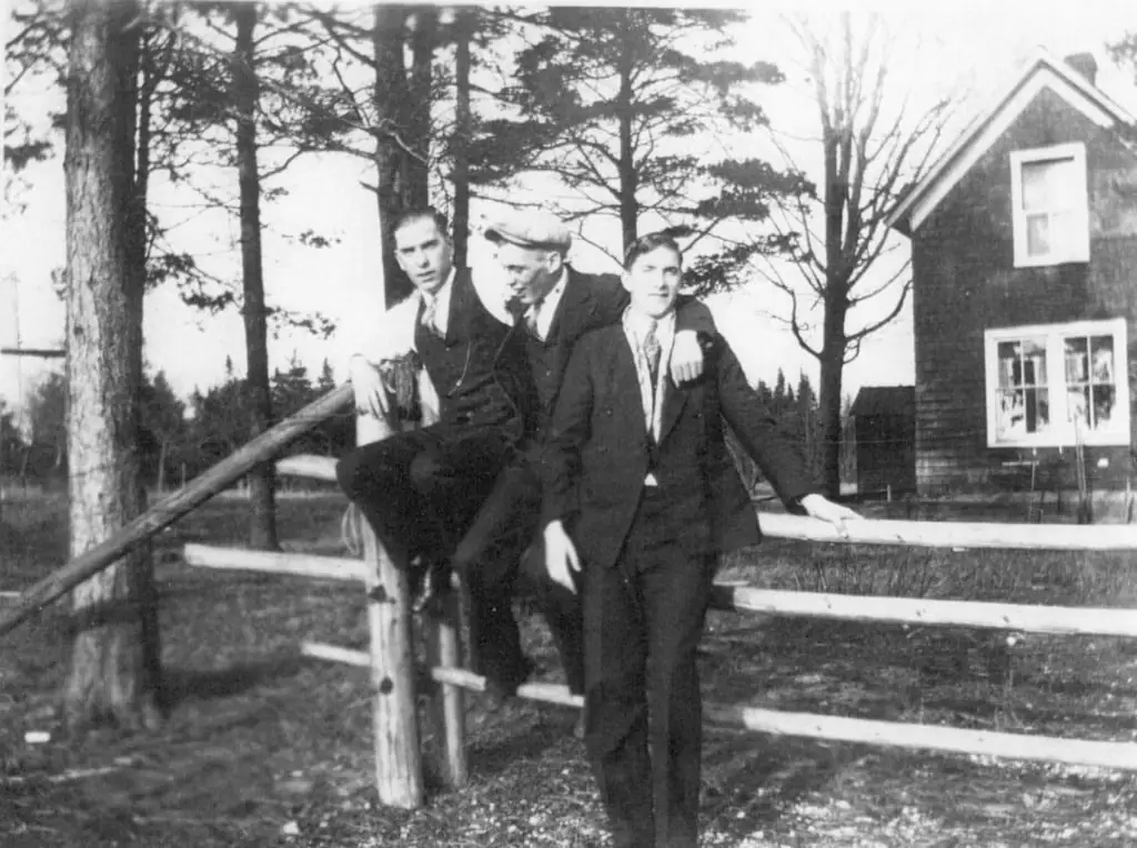 Three men in suits pose beside a wooden fence near a house and trees.