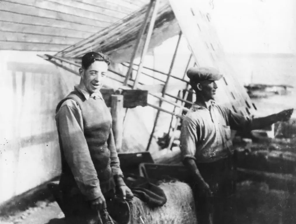 Two men stand on the deck of a sailing vessel, with one pointing toward the water.