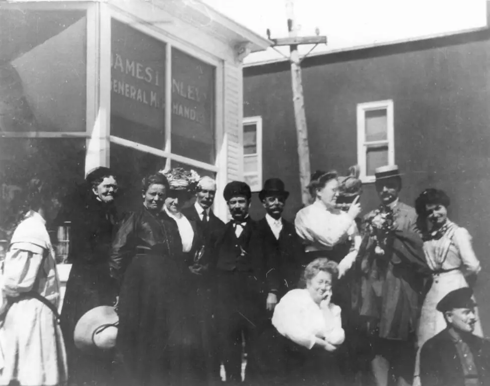 A group of adults poses outside a general store beneath painted lettering and beside a utility pole.
