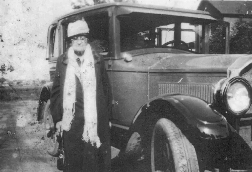 A woman in a hat and fringed scarf stands beside a large vintage automobile.