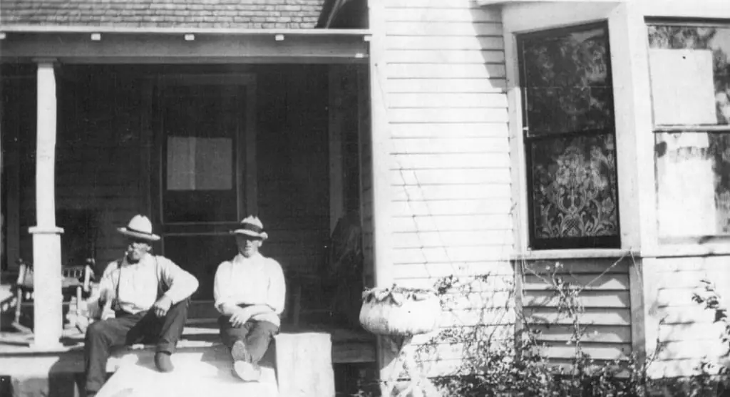 Two men in hats sit on the steps of a wooden house porch.