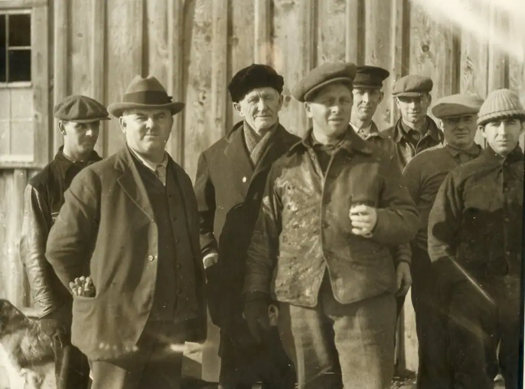 Eight men in work clothes and caps pose outside a wooden building, with a dog partly visible at lower left.