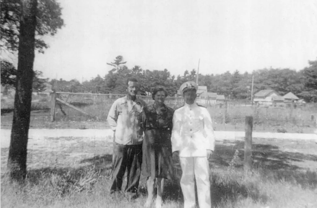Three adults stand outdoors beside a fenced grassy area, including one person wearing a white uniform and cap.