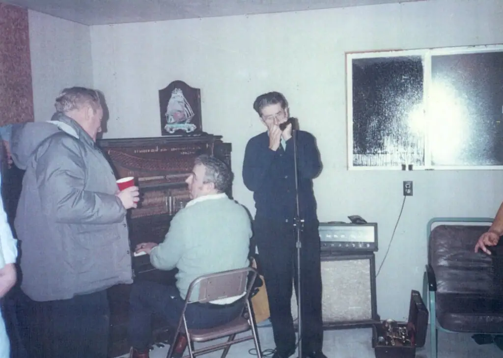 Three people gather around a piano as one sings into a microphone in a room with musical equipment.
