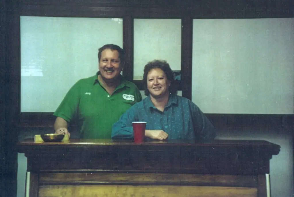 Two smiling adults stand behind a wooden counter with a red cup and small bowl.