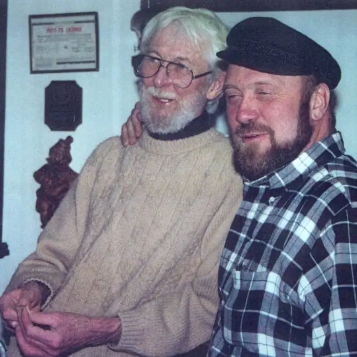 Two bearded men pose closely together indoors, one wearing glasses and a knit sweater and the other a flat cap and plaid shirt.