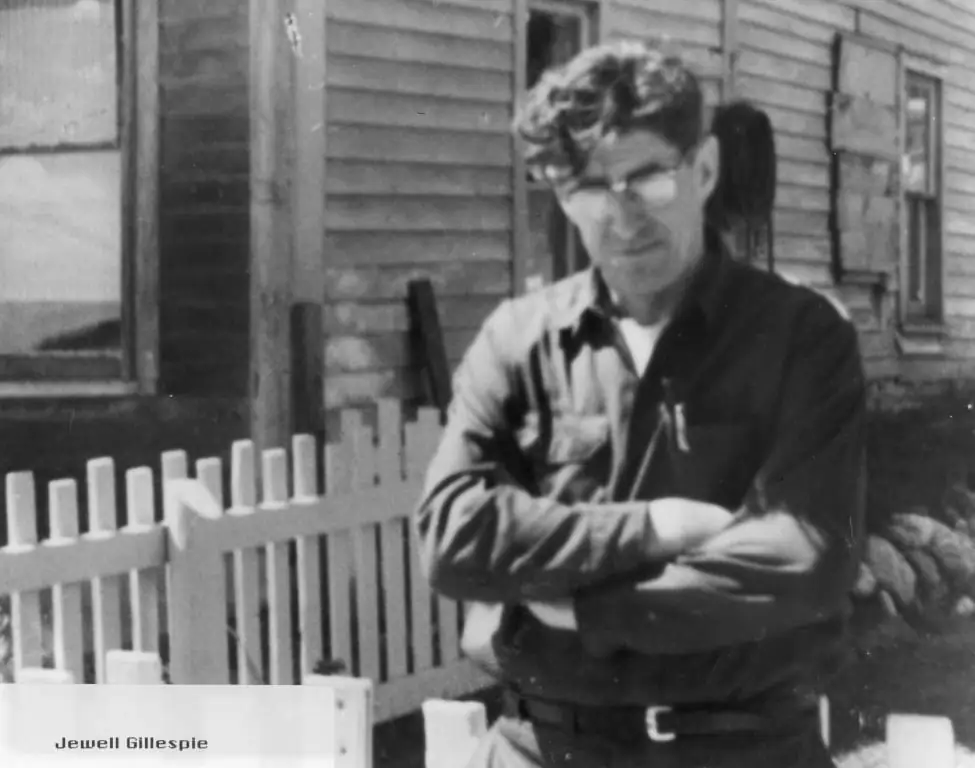 Black-and-white portrait of a man with glasses standing with folded arms beside a picket fence.