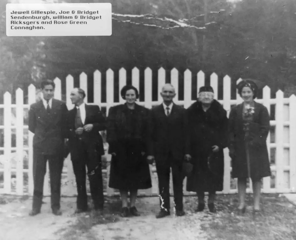 Six adults in formal clothing stand in front of a white picket fence, with one woman holding a small bouquet.
