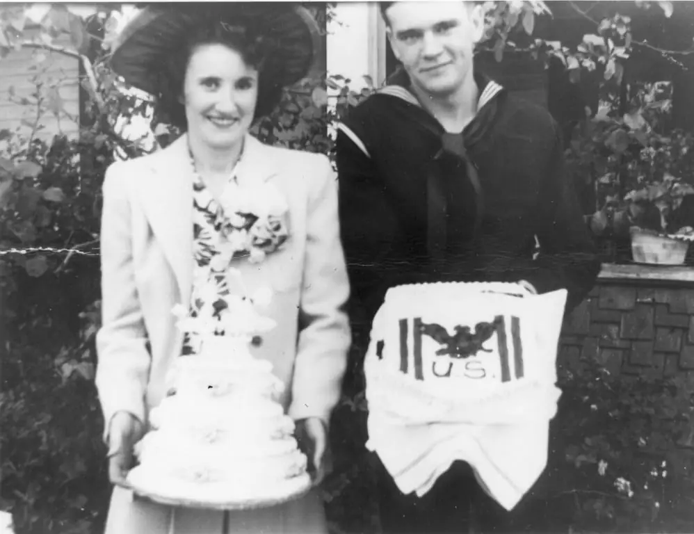 A bride holding a tiered wedding cake stands beside a groom in a sailor uniform.