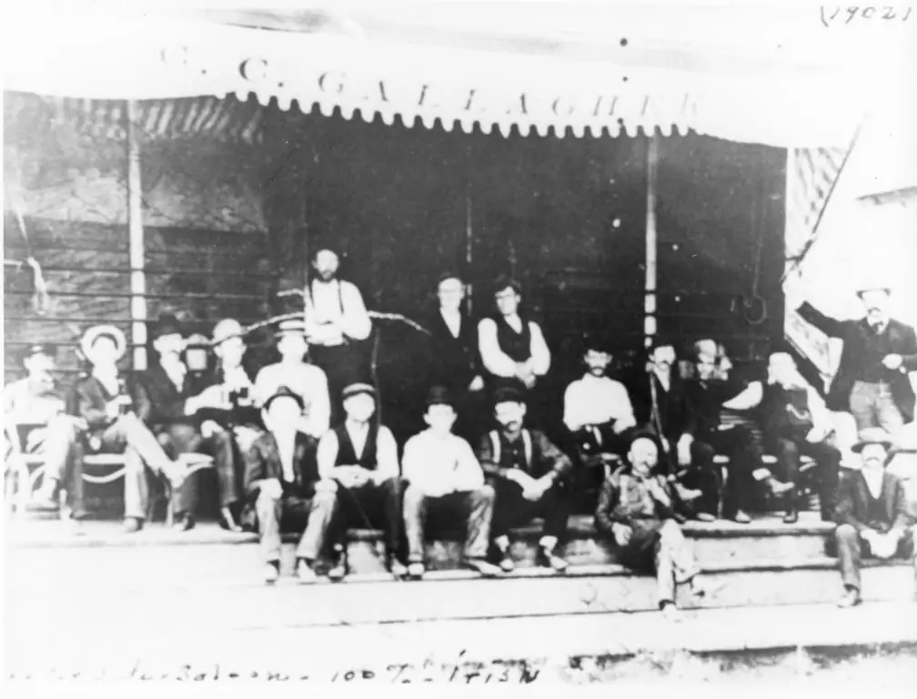 A group of men pose on the steps of a building marked “C. C. GALLAGHER.”