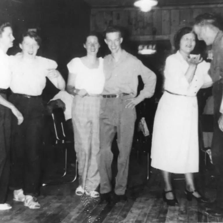 Six adults gather indoors, with one pair facing each other as if dancing.