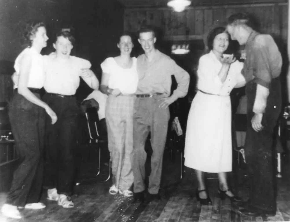 Six young adults pose together in a wood-paneled room with chairs and a wooden floor.