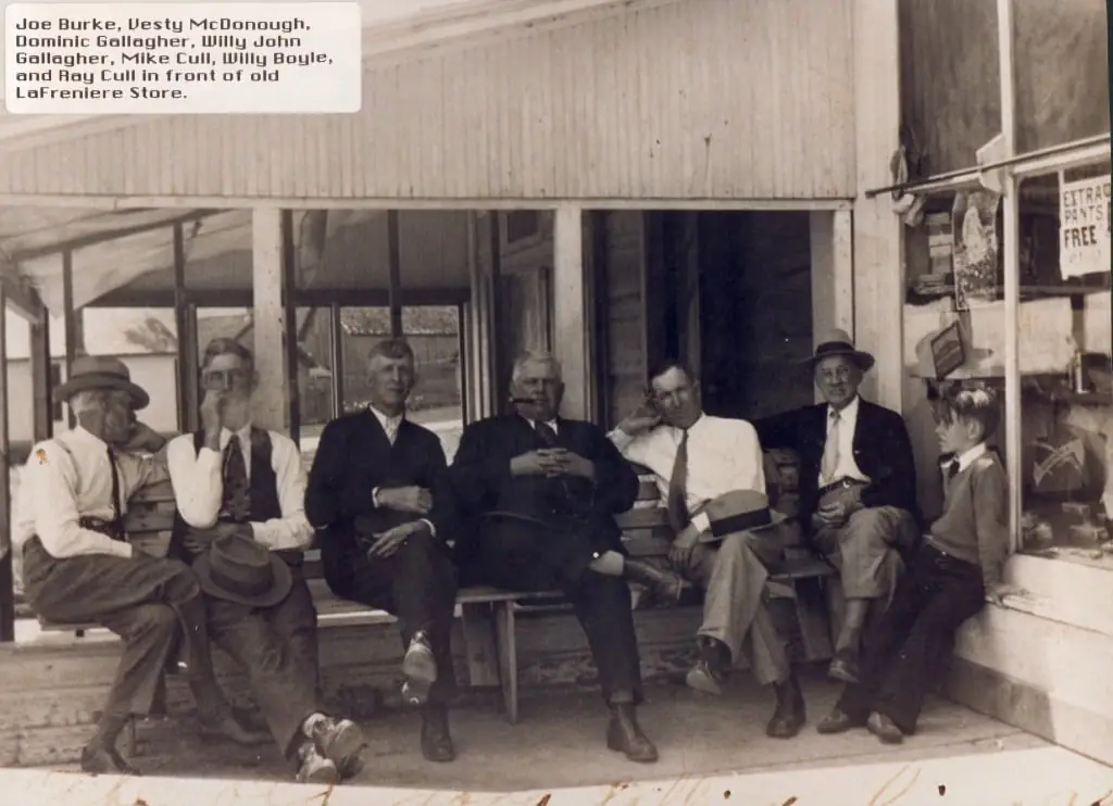 A group of men and a boy sit on a bench outside a wooden storefront.