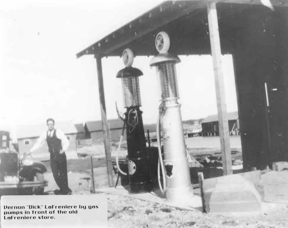 A man stands beside two vintage gas pumps and an old automobile under a wooden canopy.