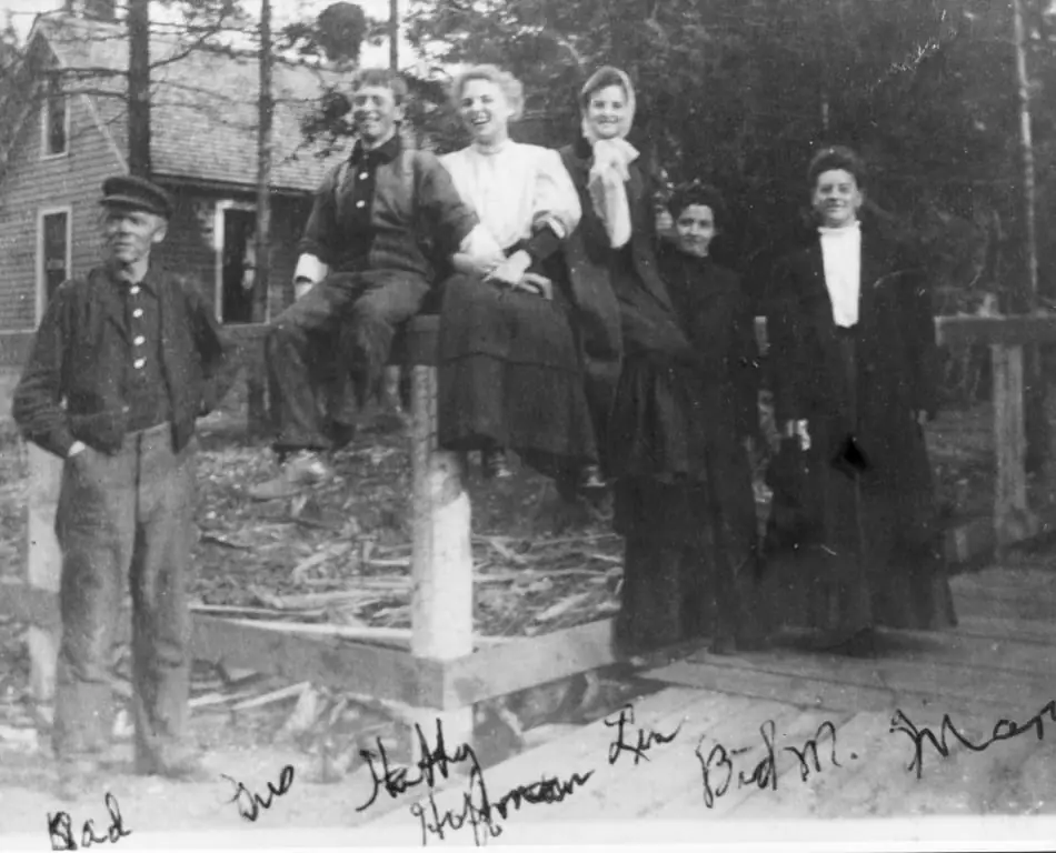 Six people pose beside a wooden house, with handwritten names along the bottom of the photograph.