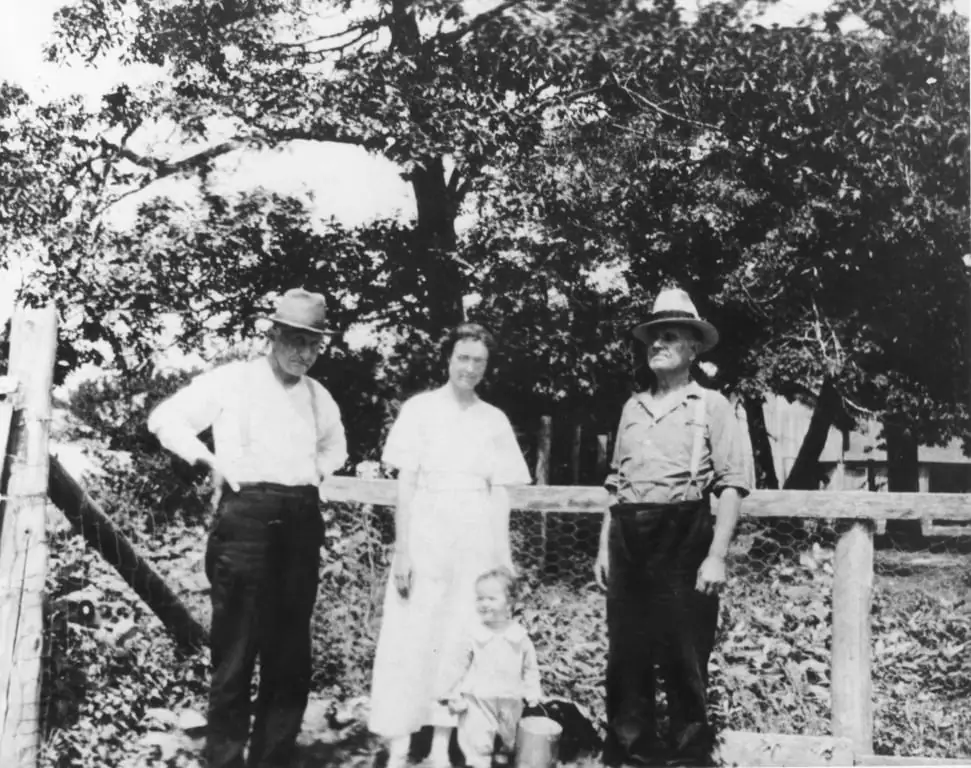 Four people, including a child, pose beside a wooden and wire fence beneath large trees.
