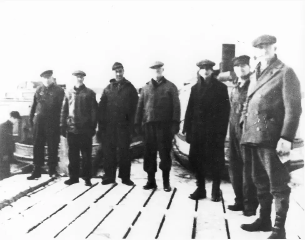 Seven men in heavy coats and caps stand on a snow-covered wooden pier beside a partially visible vessel.