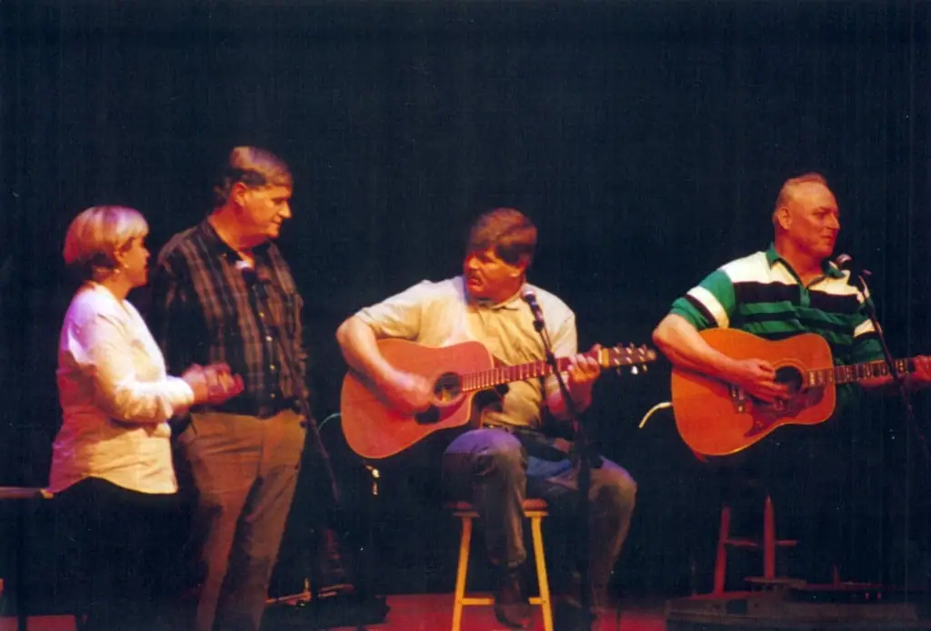 Four musicians perform on a dimly lit stage, with two acoustic guitars and microphones.