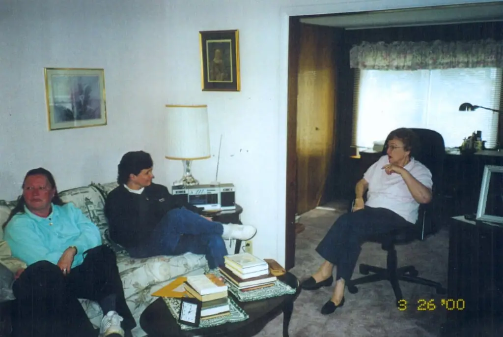 Three women sit in a furnished living room with books on a coffee table.