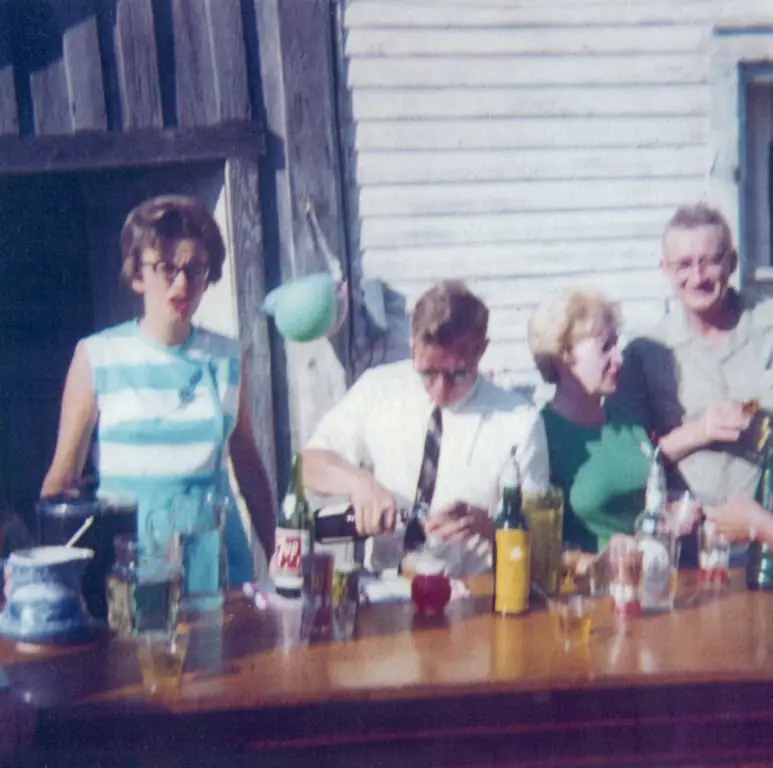 Four adults gather behind a bar, with one person pouring a drink among bottles and glasses.