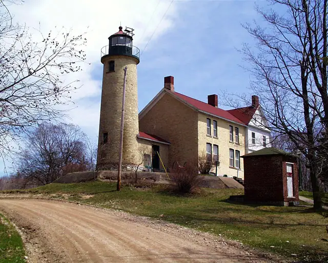 A stone lighthouse and adjoining two-story residence stand beside a dirt road among leafless trees.