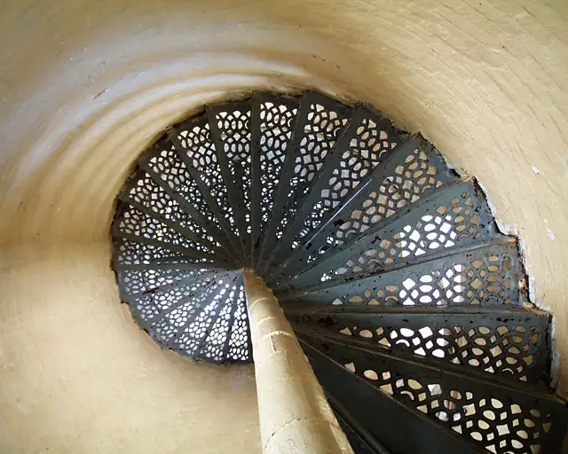 Top-down view of a perforated metal spiral staircase inside a round, light-colored shaft.
