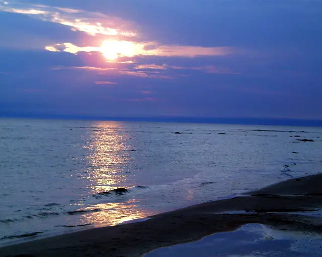 Sunset light reflects across calm water beside a dark shoreline and shallow foreground pool.