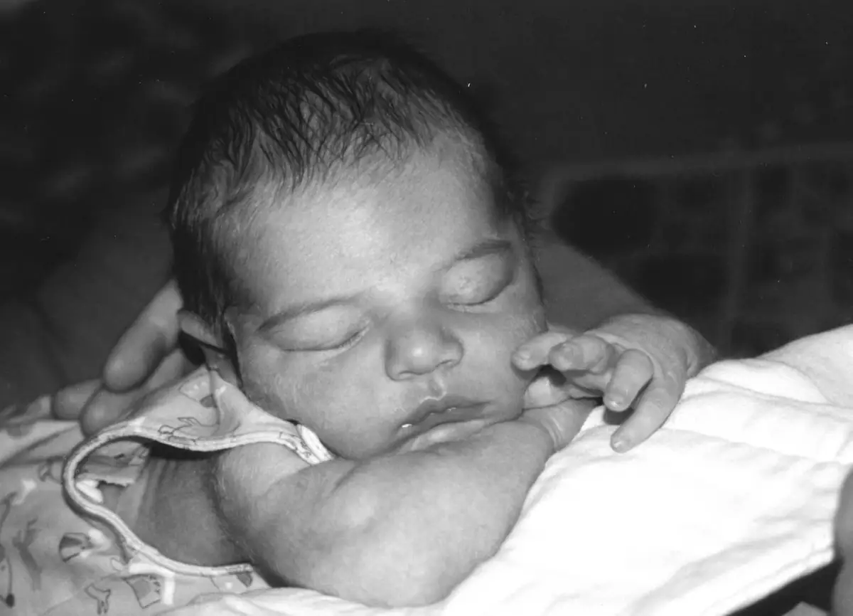 Sleeping infant resting on a patterned garment and white blanket, with hands supporting the head and face.