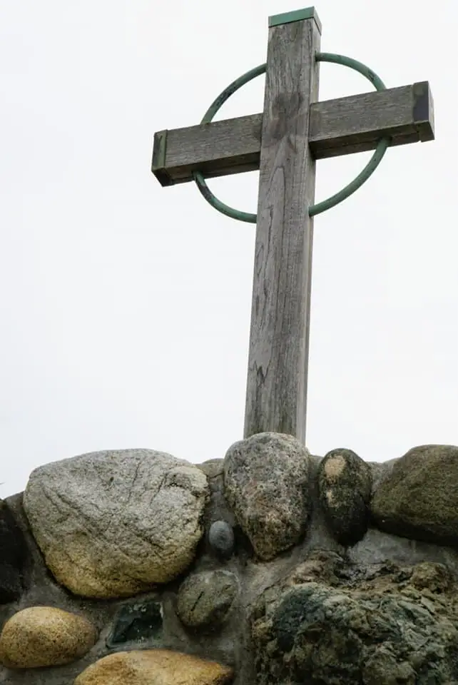 A weathered wooden cross with a circular metal ring stands among large rounded stones beneath an overcast sky.