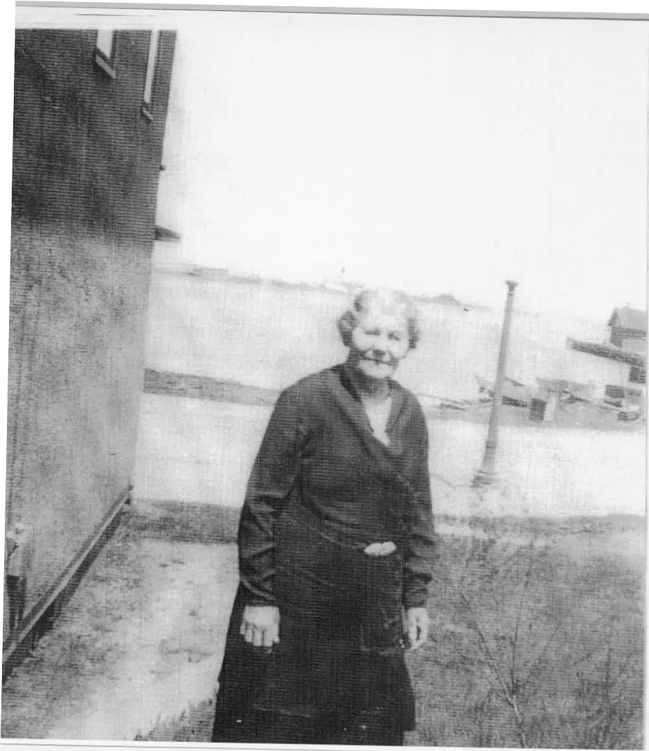Black-and-white portrait of an elderly woman standing beside a waterfront building.