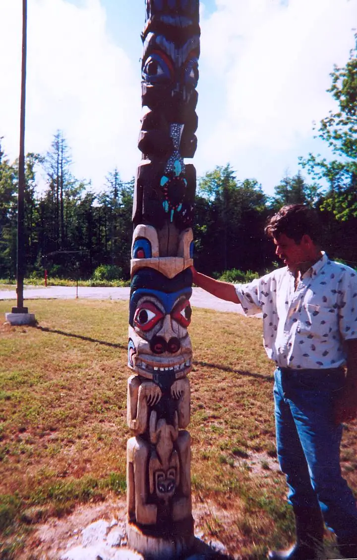 A man stands beside a tall, brightly painted wooden carved pole outdoors.