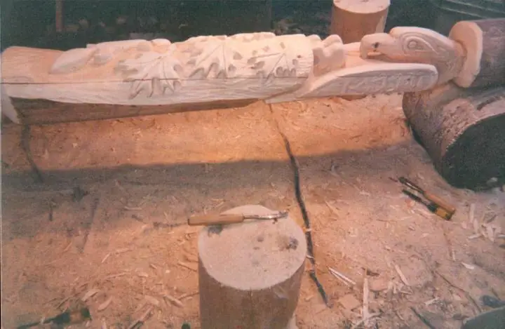 A partially carved wooden sculpture rests on logs above a sawdust-covered work area.