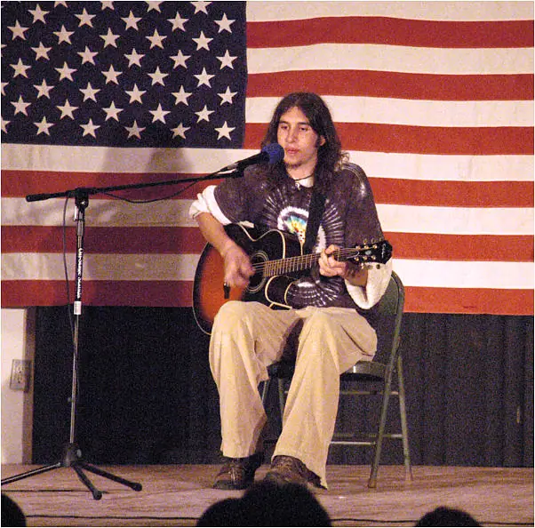 Long-haired performer seated onstage, playing an acoustic guitar before a large American flag.