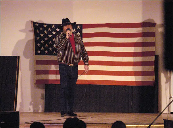 A performer in a cowboy hat stands at a microphone before a large American flag.
