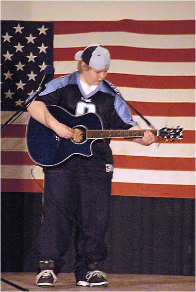 A performer in a cap and sports jersey plays an acoustic guitar before a large American flag.