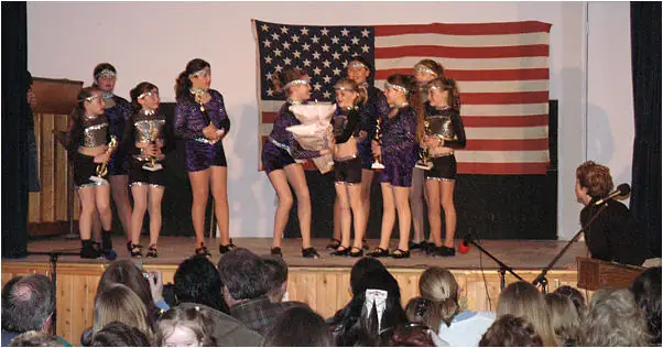 Young performers stand on a stage before an American flag while an audience watches.