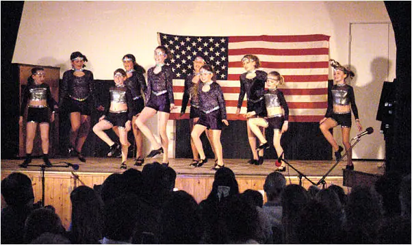Nine dancers perform on a stage before an American flag while an audience watches.