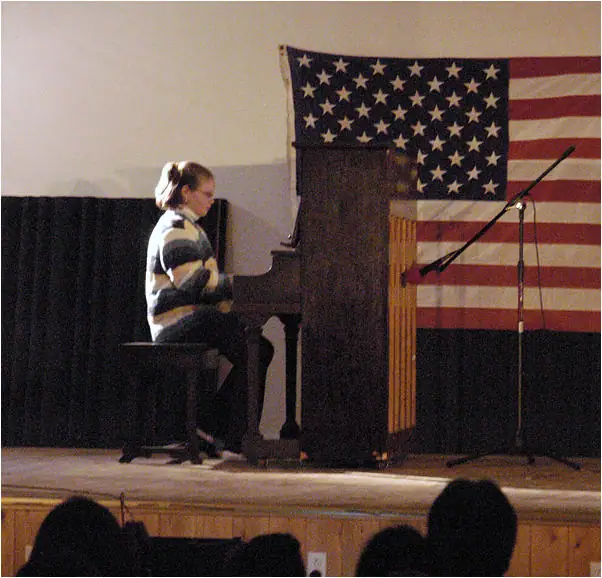 A pianist performs onstage at a grand piano before an audience, with a large flag behind her.